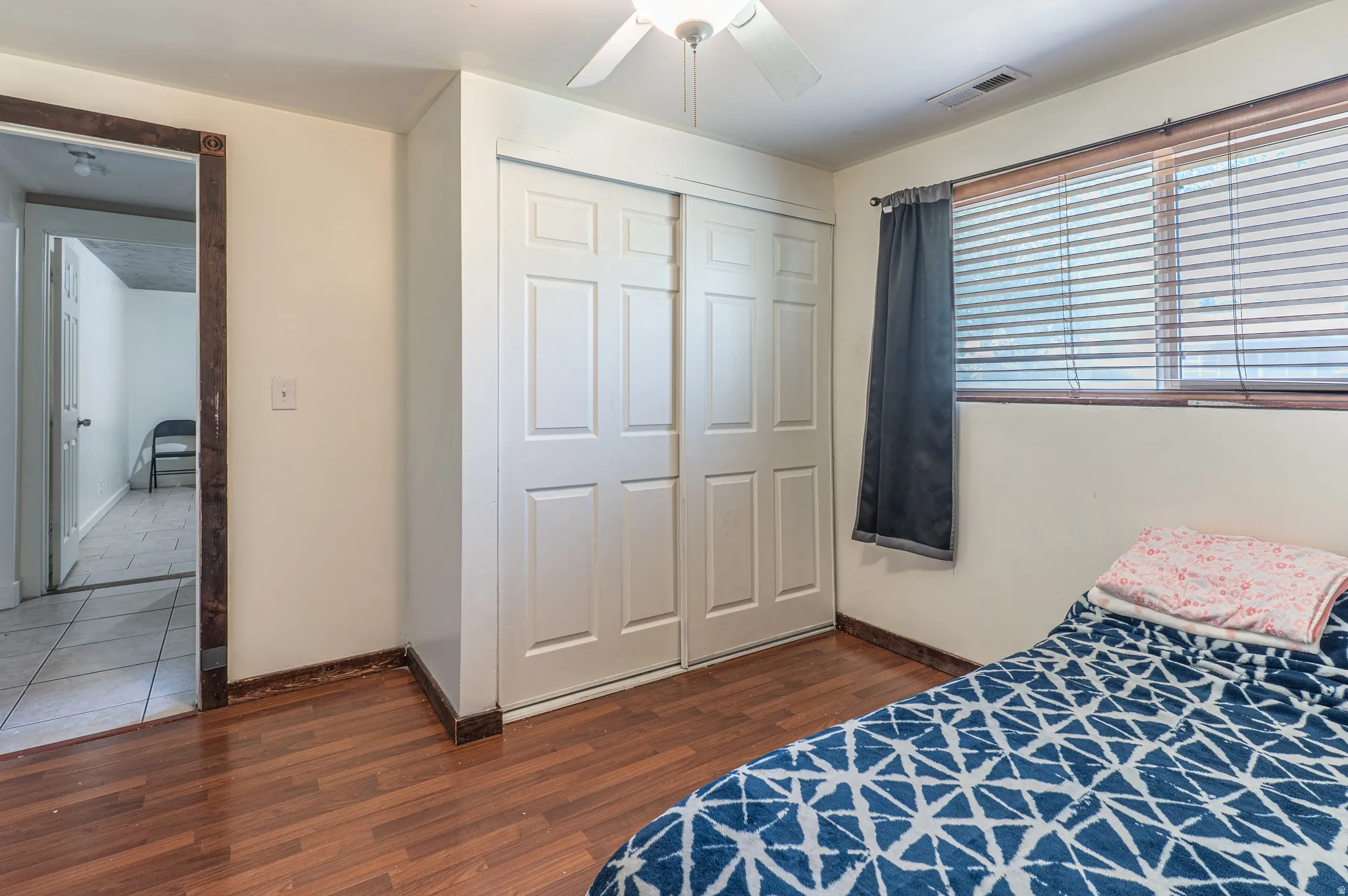 Room featuring wood-finish flooring, a ceiling fan, window with blinds, and white bi-fold closet doors
