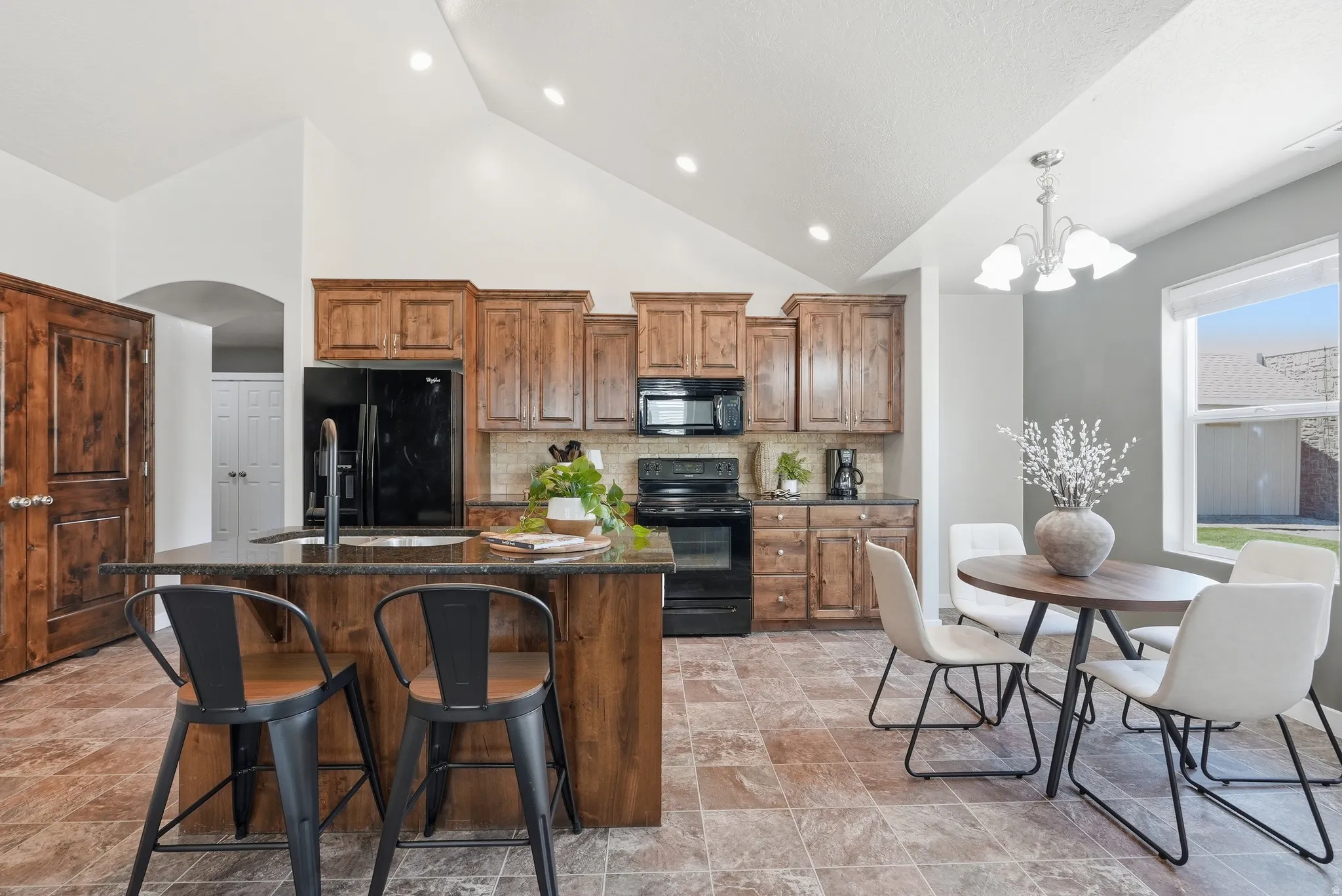 Kitchen featuring wood cabinetry, black appliances, a central island with a sink, vaulted ceilings, and abundant natural light