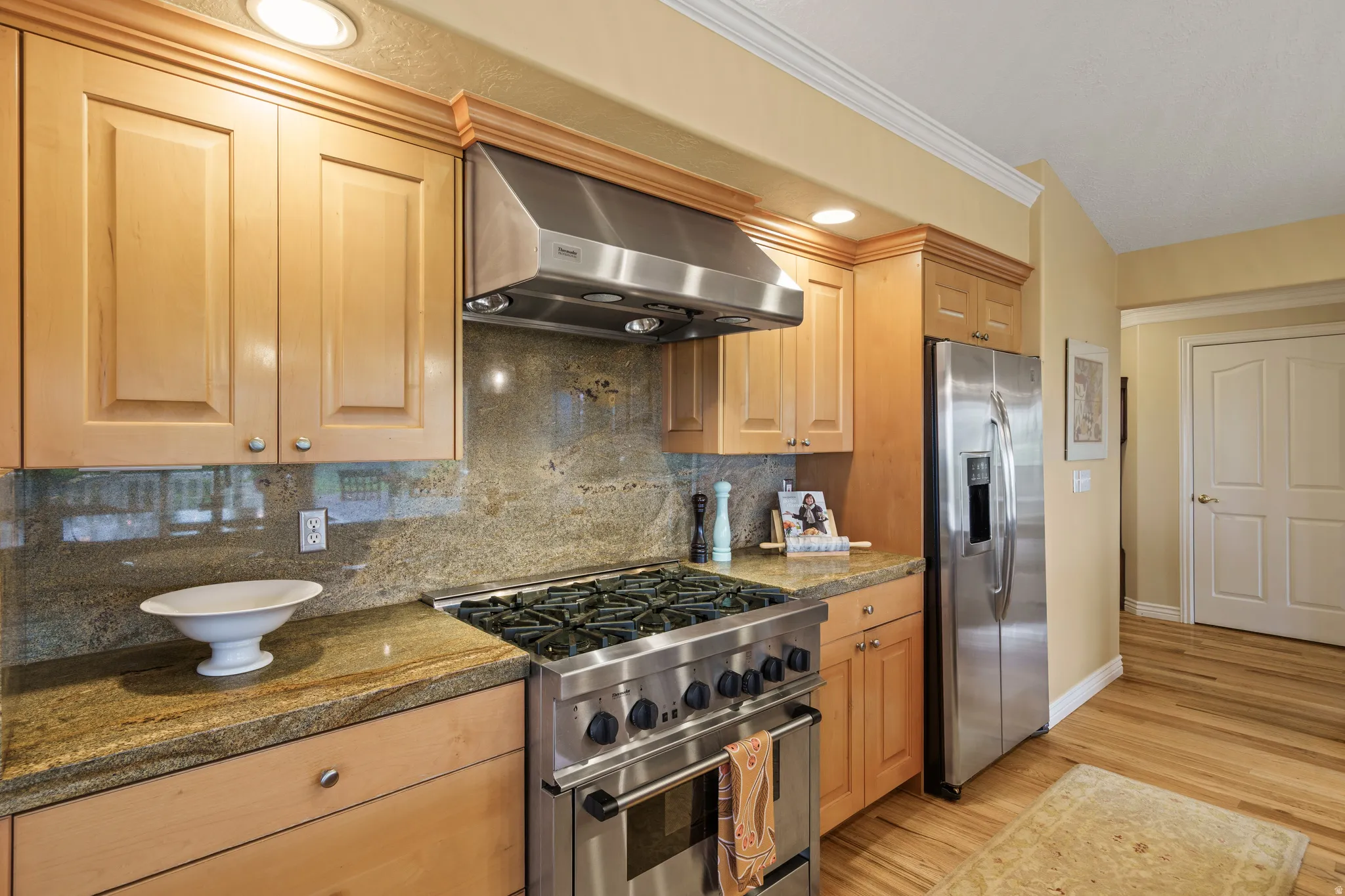 Gourmet kitchen featuring a professional Thermador cooktop  stainless steel range hood, stone countertops, light wood-finish cabinetry, and a full-size stainless steel refrigerator