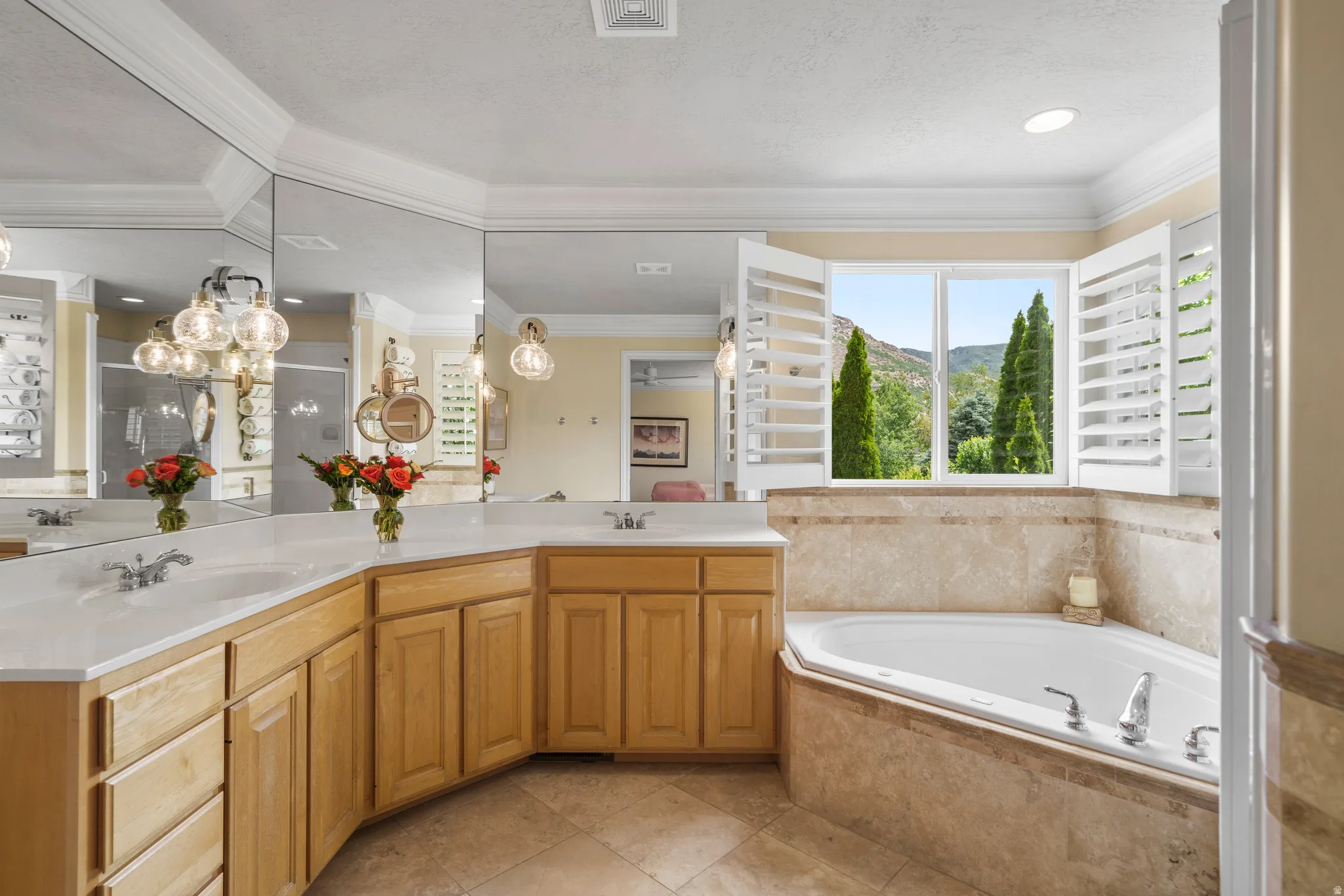 Primary Bathroom featuring a built-in corner soaking tub with tiled surround, a large window with white plantation shutters, mountain views  and a vanity with dual sinks, light wood cabinetry, and extensive mirror coverage