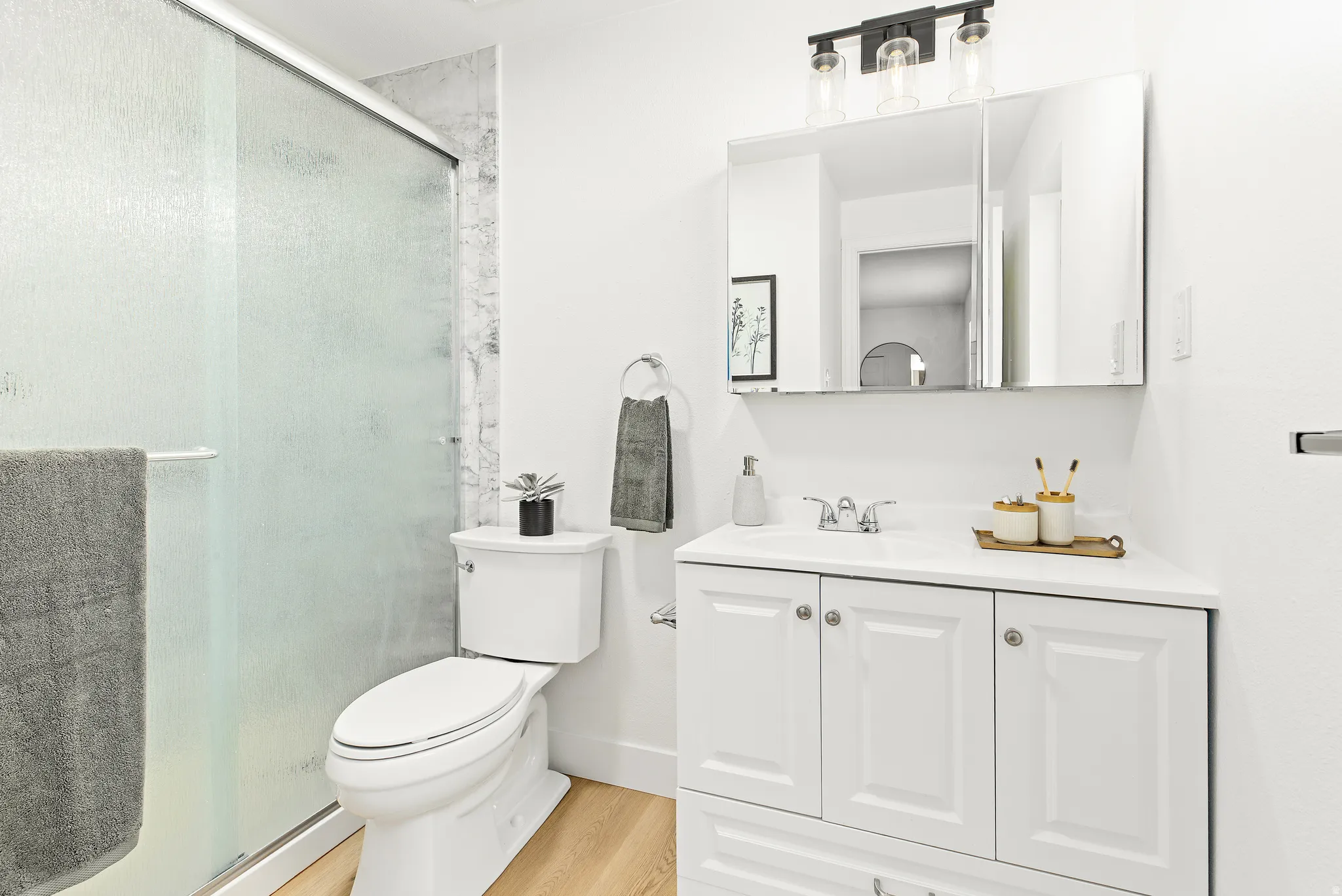 Bathroom featuring a white vanity with panel cabinetry and a white countertop, a mirrored medicine cabinet, a three-light vanity fixture, a toilet, and a shower with a frosted glass enclosure and marble-look tile surround