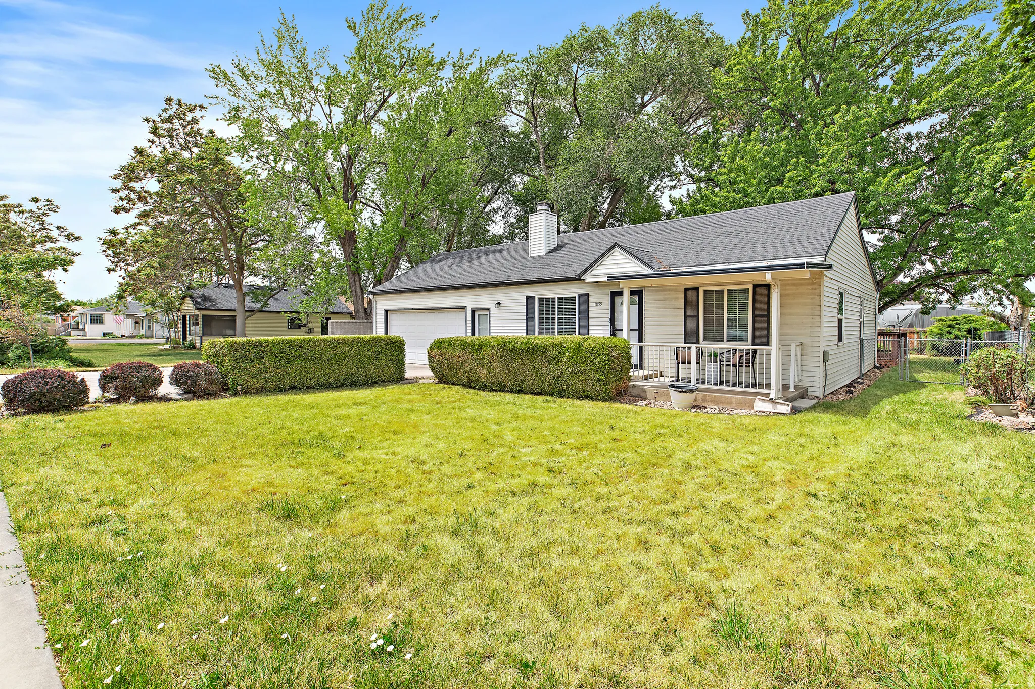 Single-story residence featuring light-colored siding, a dark shingle roof, a front porch with white railings, a front-facing garage, and mature landscaping