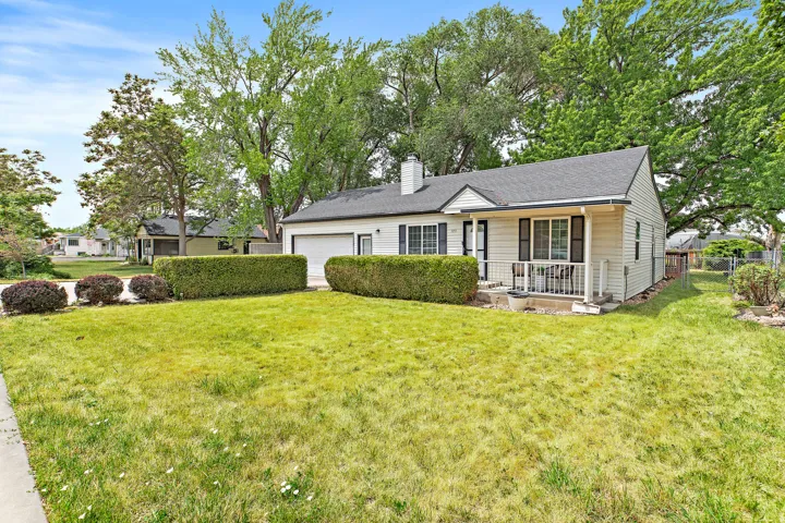 Single-story residence featuring light-colored siding, a dark shingle roof, a front porch with white railings, a front-facing garage, and mature landscaping