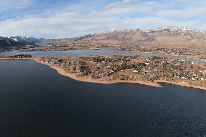 Expansive aerial showcasing a large body of water, a residential area with varied housing styles, surrounding natural landscapes, and distant snow-capped mountains