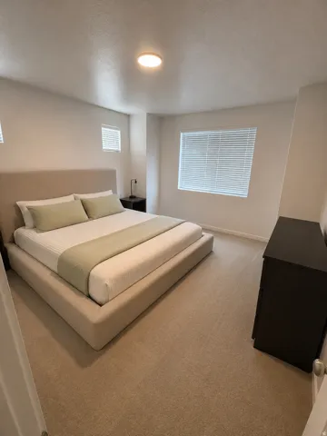 Spacious bedroom featuring neutral carpeting, white walls, multiple windows with blinds, and a flush-mount ceiling light fixture