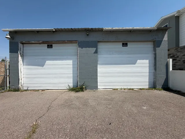 Two white roll-up garage doors set into a gray concrete block structure, featuring an asphalt driveway and an overhead eave