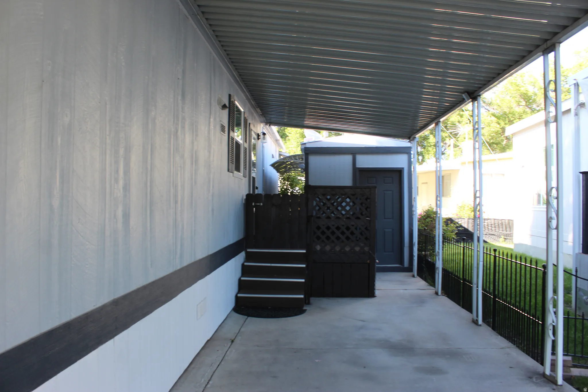 Covered carport and side entry featuring a privacy lattice, dark-painted steps, and a black wrought iron fence. Leads into the mudroom or laundry room.  Side door has a storm door too.
