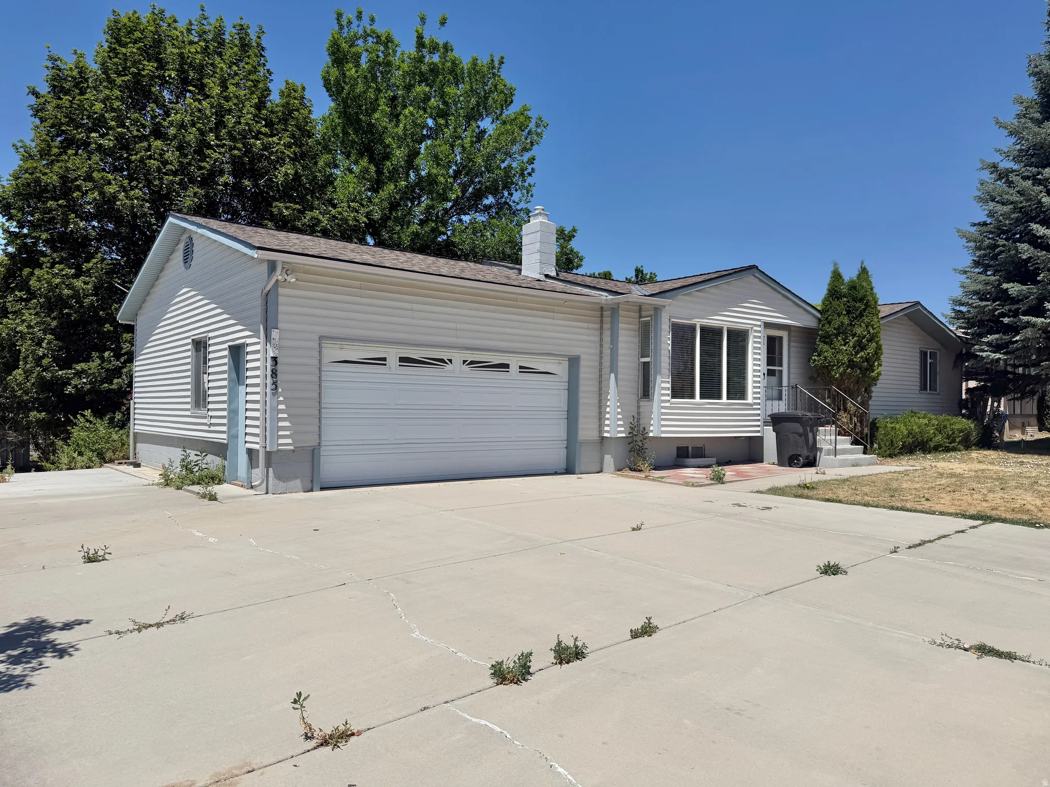 Exterior featuring light-colored siding, a two-car garage, a concrete driveway, and mature trees