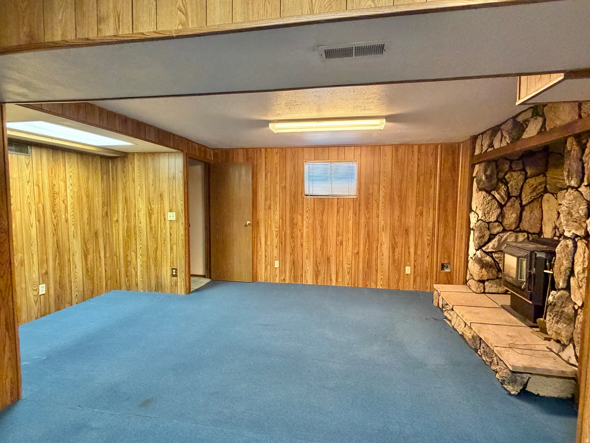 Spacious room featuring wood-finish wall paneling, blue carpet, a stone-clad fireplace with hearth, and a window