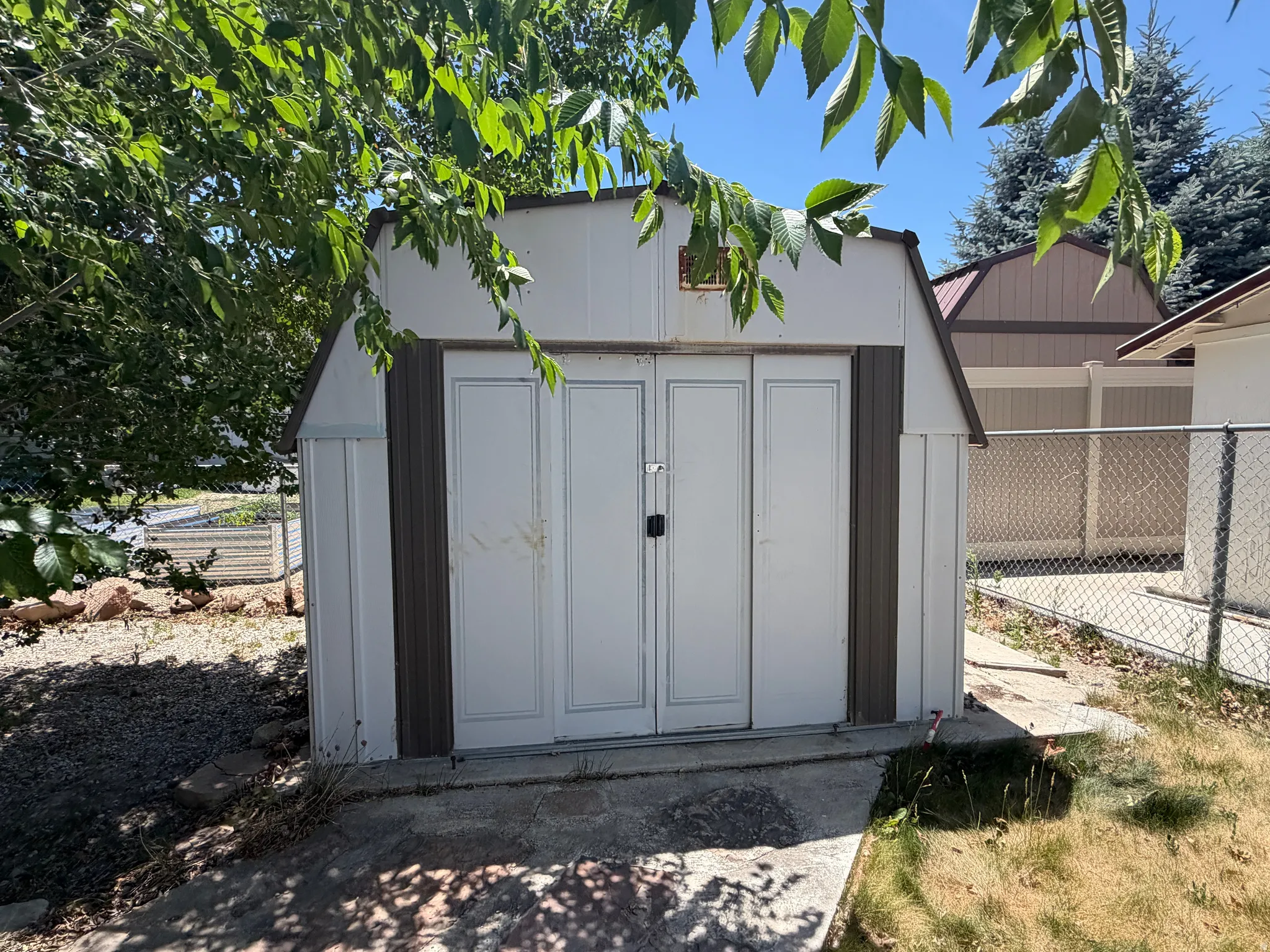 White and brown exterior shed with double doors, concrete foundation, and chain-link fencing