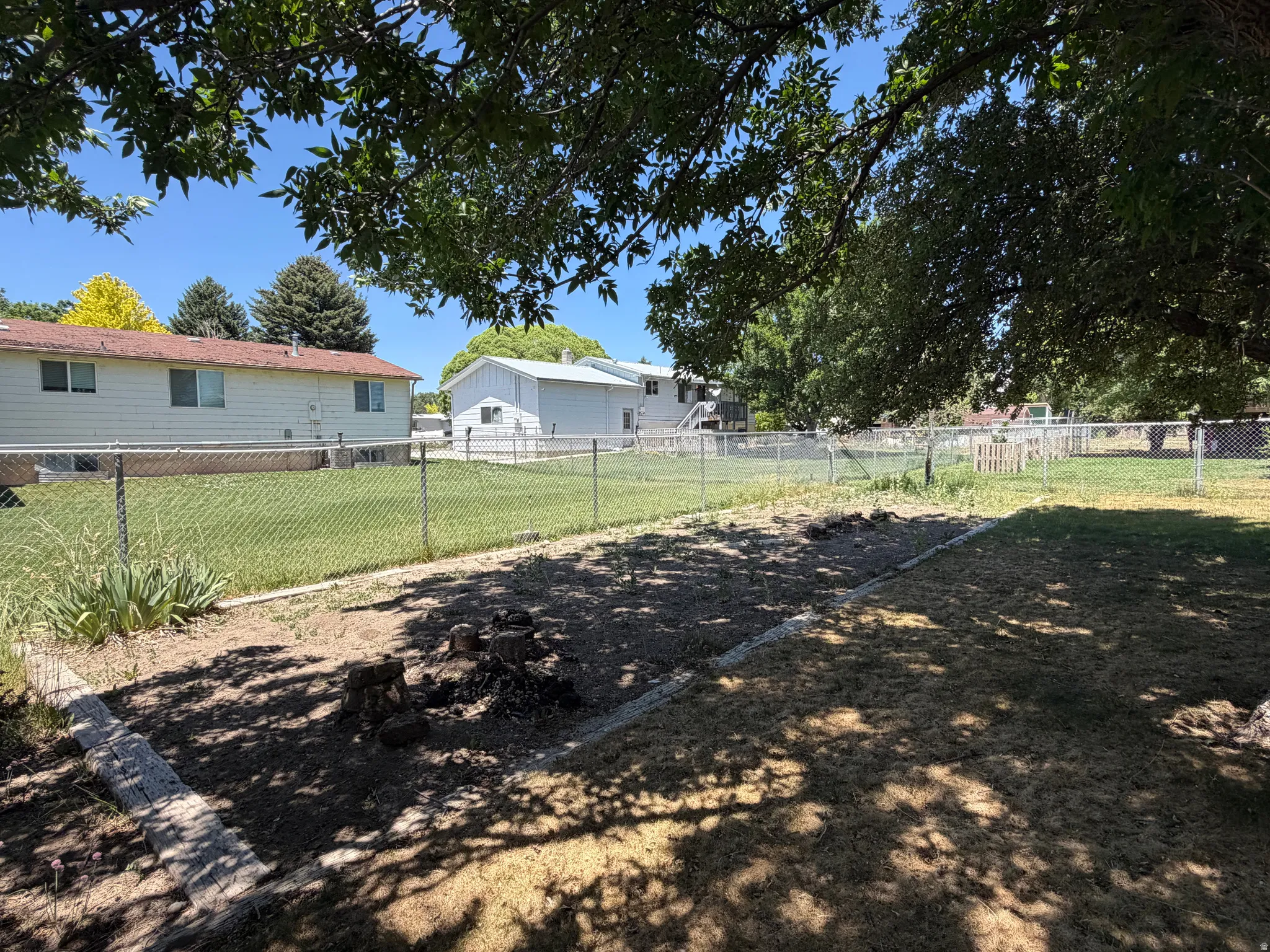 Expansive backyard featuring a partial chain-link fence, mature trees providing shade, and a designated garden bed