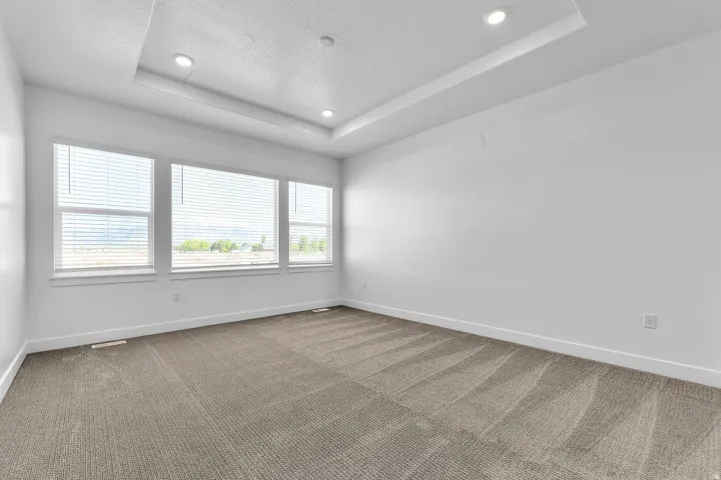 Spacious room featuring a tray ceiling with recessed lighting, three large windows, textured carpet flooring, and white baseboards