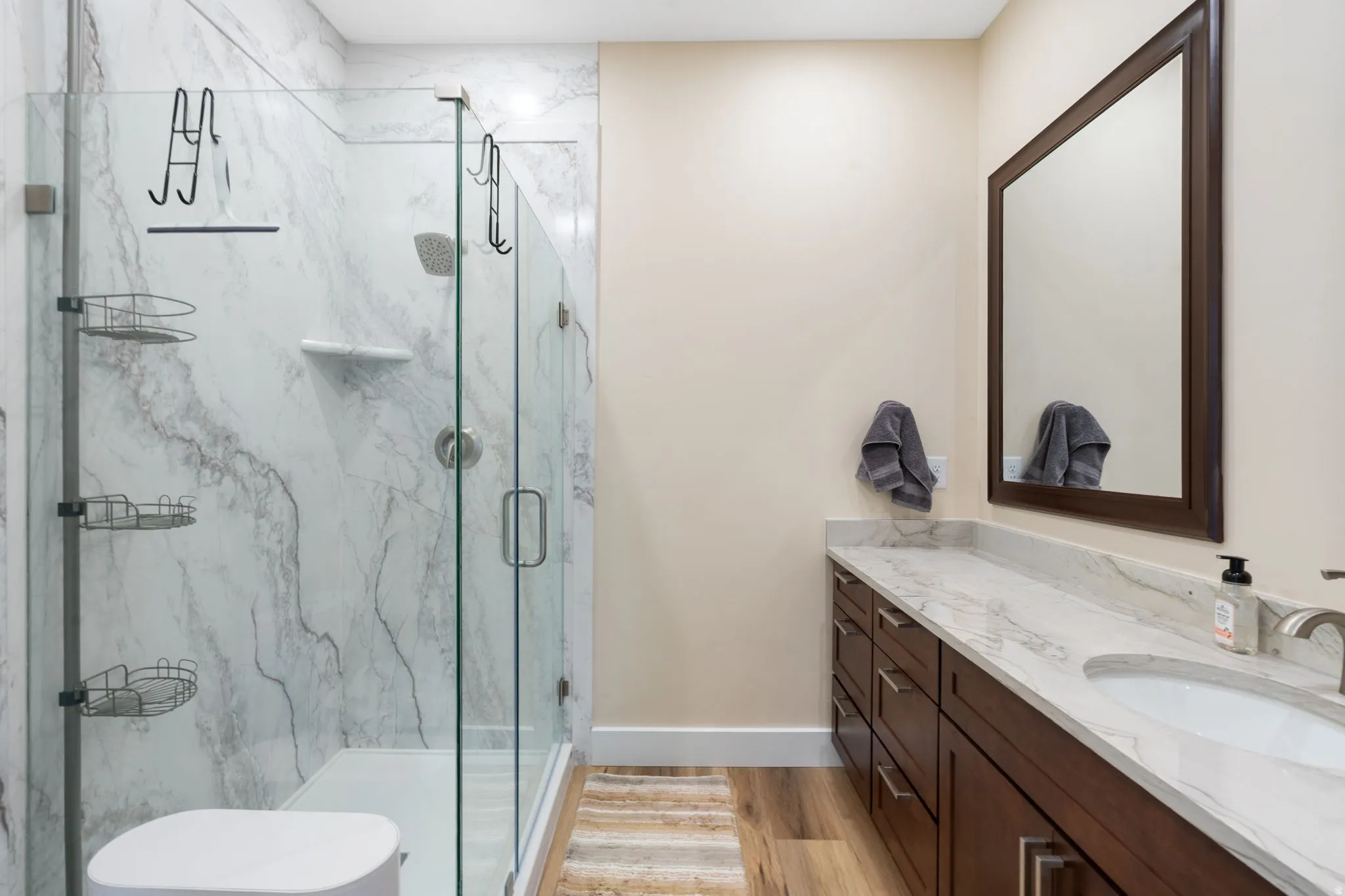 Spacious bathroom featuring a glass-enclosed shower with marble-look wall panels, a dark wood-finish vanity, a light-toned countertop, and wood-finish flooring