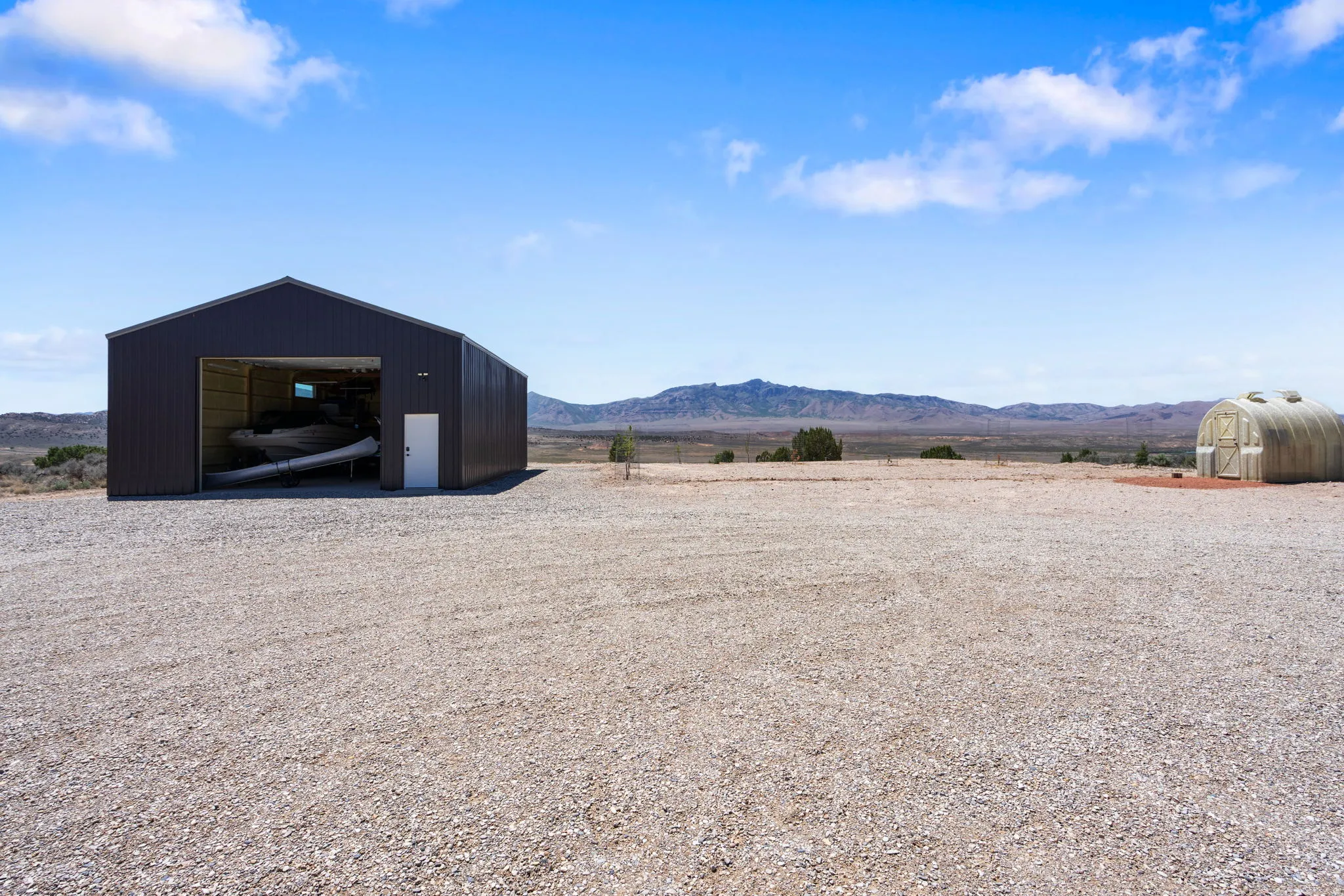 Expansive gravel lot featuring a large metal building with a roll-up door and a separate entry door, alongside a hoop house
