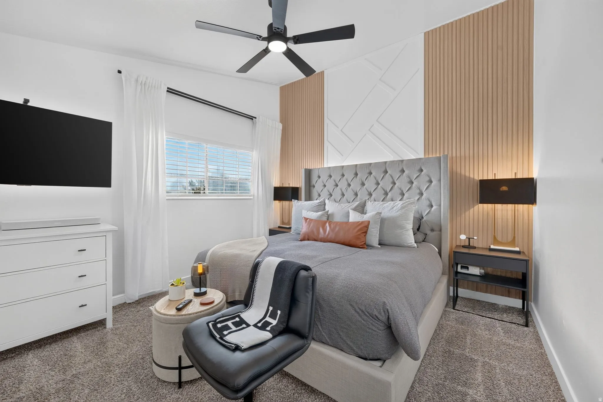 Accent wall with geometric and vertical paneling, plush carpet flooring, a ceiling fan with integrated lighting, a window with blinds and drapes, and a built-in wall-mounted television