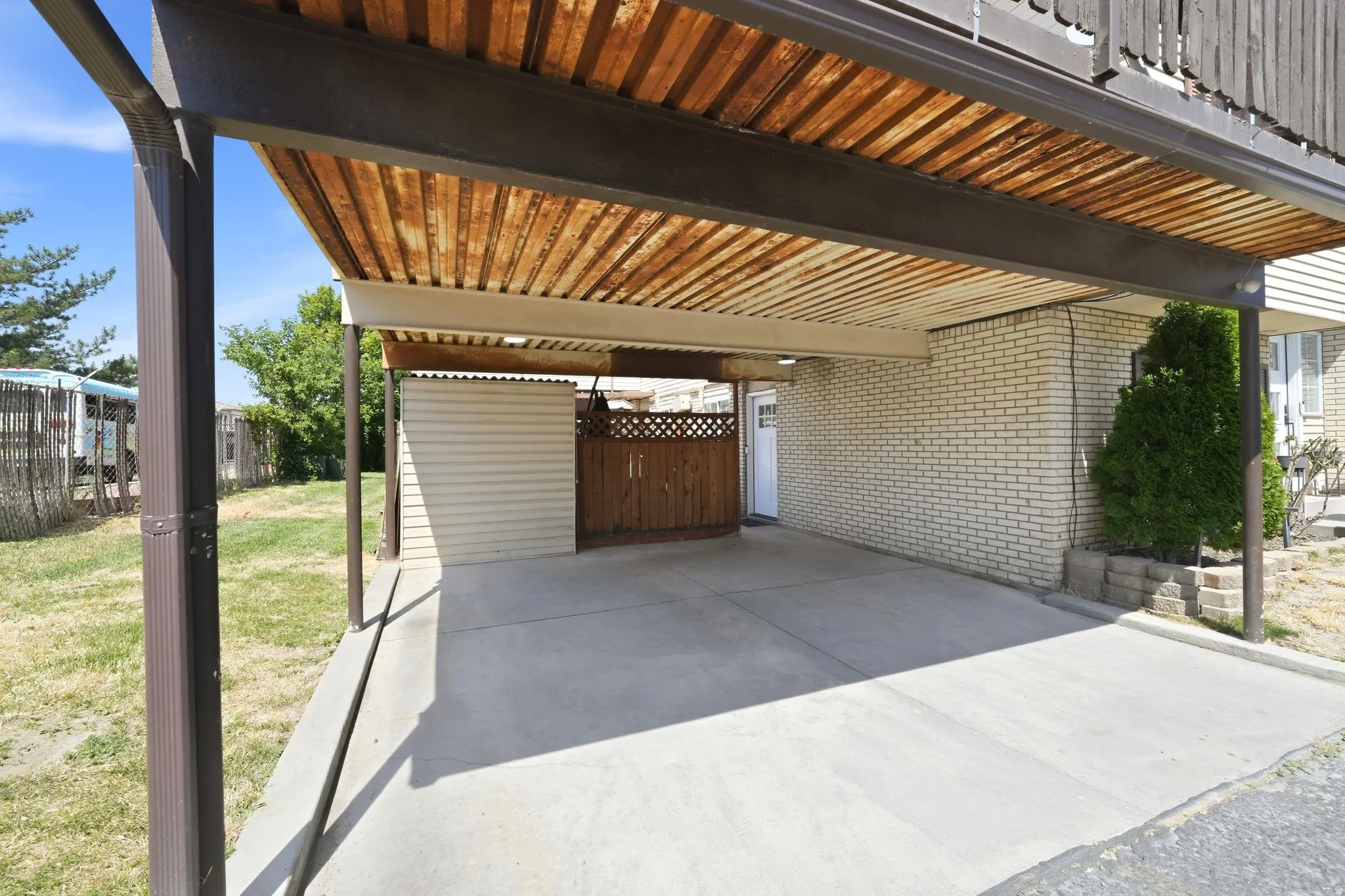 Concrete carport featuring a slatted wood ceiling, a light brick exterior wall, and a built-in storage shed with a coordinating wood privacy fence