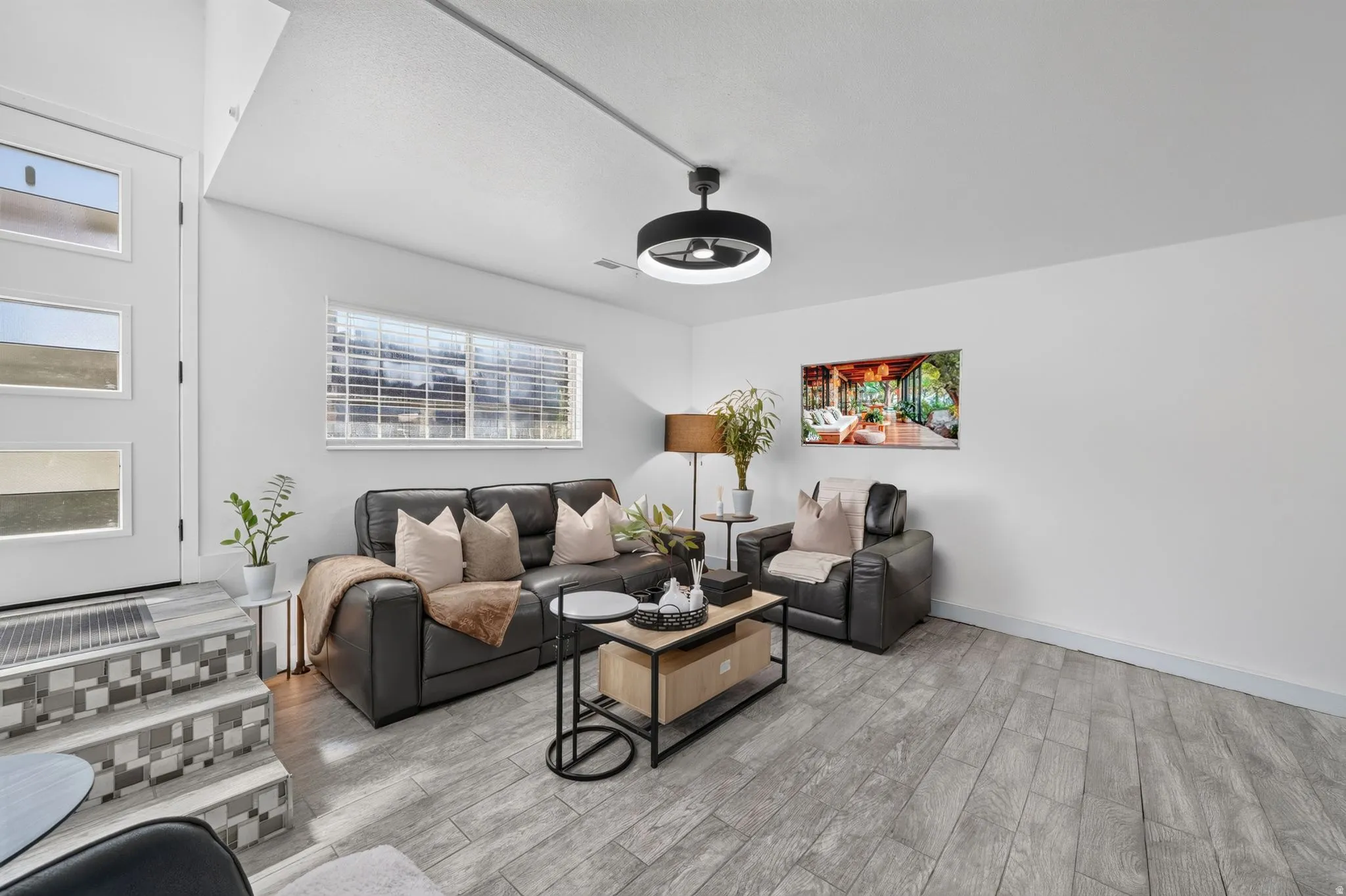 Spacious living area featuring wood-finish flooring, a contemporary ceiling fan with integrated lighting, and a large window with blinds