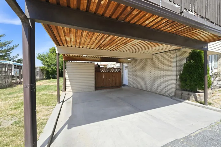 Concrete carport featuring a slatted wood ceiling, a light brick exterior wall, and a built-in storage shed with a coordinating wood privacy fence