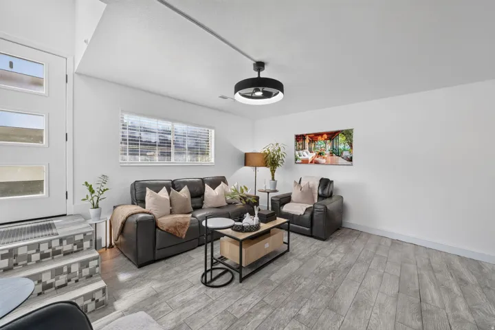 Spacious living area featuring wood-finish flooring, a contemporary ceiling fan with integrated lighting, and a large window with blinds
