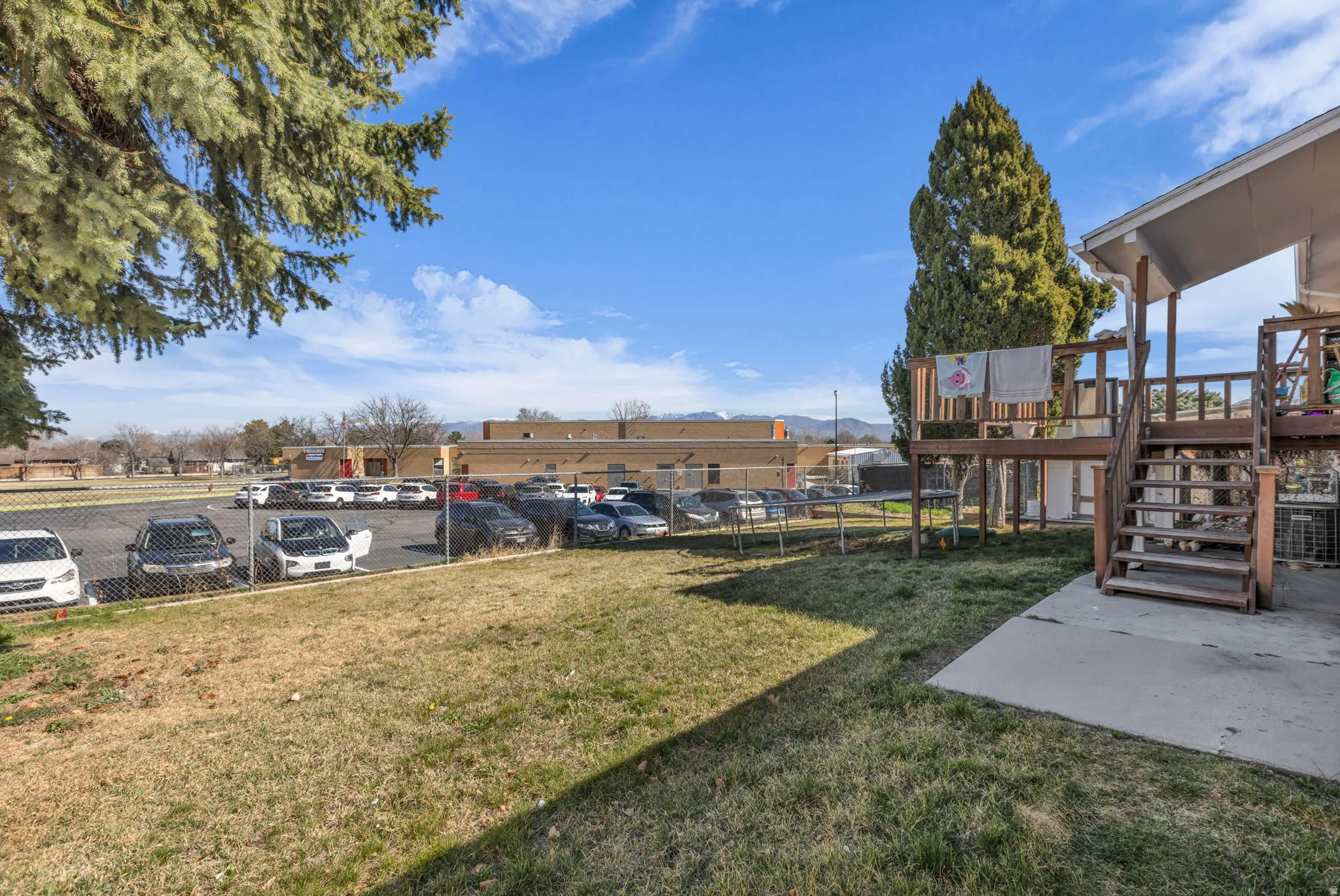 Spacious backyard featuring a wooden deck with railing and stairs, mature trees, and a concrete patio area