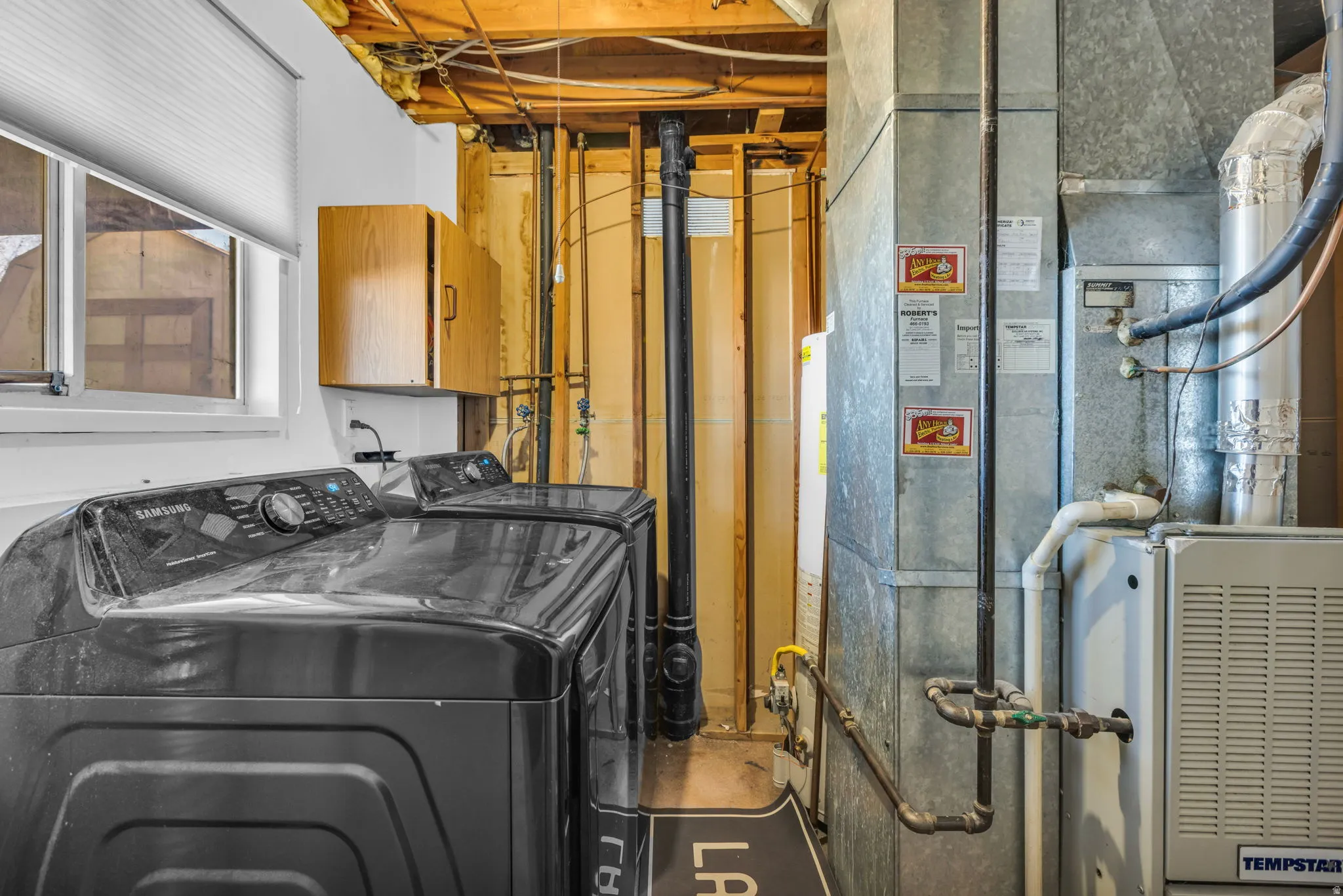 Utility room featuring exposed ceiling joists, durable concrete flooring, a double window with blinds, and built-in wood cabinetry for storage