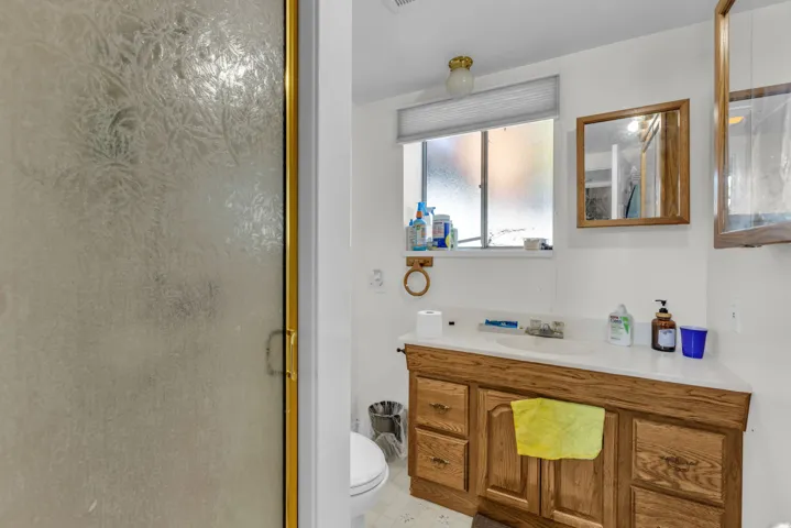 Bathroom featuring a frosted glass shower enclosure with gold-tone trim, a wood-finish vanity with a built-in sink, a wall-mounted mirror with a wood frame, and a window with horizontal blinds