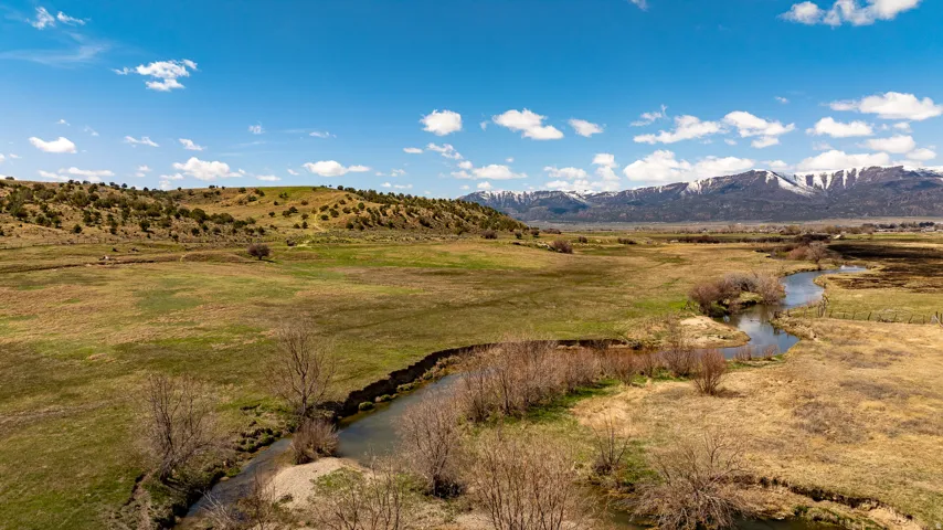 Expansive natural landscape featuring a meandering creek, diverse terrain with green and golden grasses, and distant snow-capped mountains