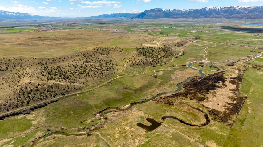 Expansive rural landscape featuring a winding river, verdant fields, rolling hills, and distant snow-capped mountains