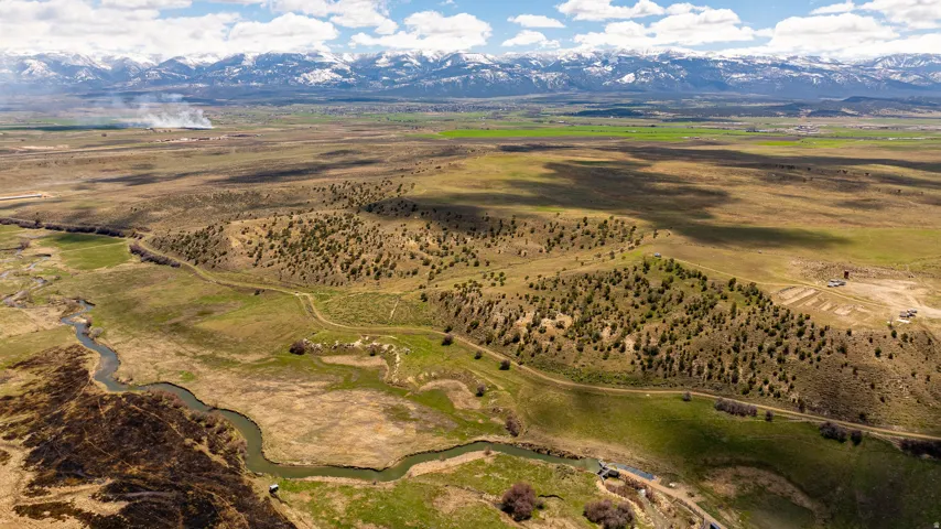 Expansive acreage featuring diverse terrain, a winding river, verdant fields, scattered trees, and a distant snow-capped mountain range