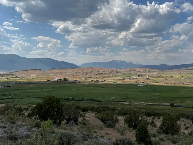 Expansive landscape featuring distant mountains, a wide valley with green fields, and scattered structures under a partly cloudy sky