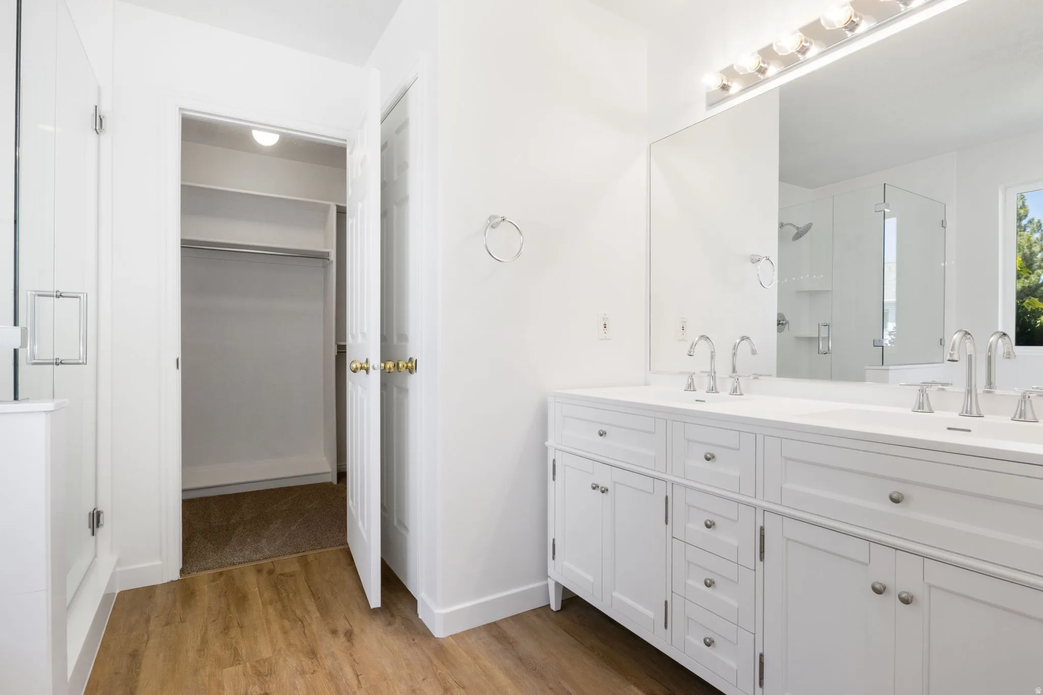 Bathroom featuring a dual vanity with white cabinetry and integrated sinks, a large mirror with strip lighting, wood-finish flooring, and a glass-enclosed shower