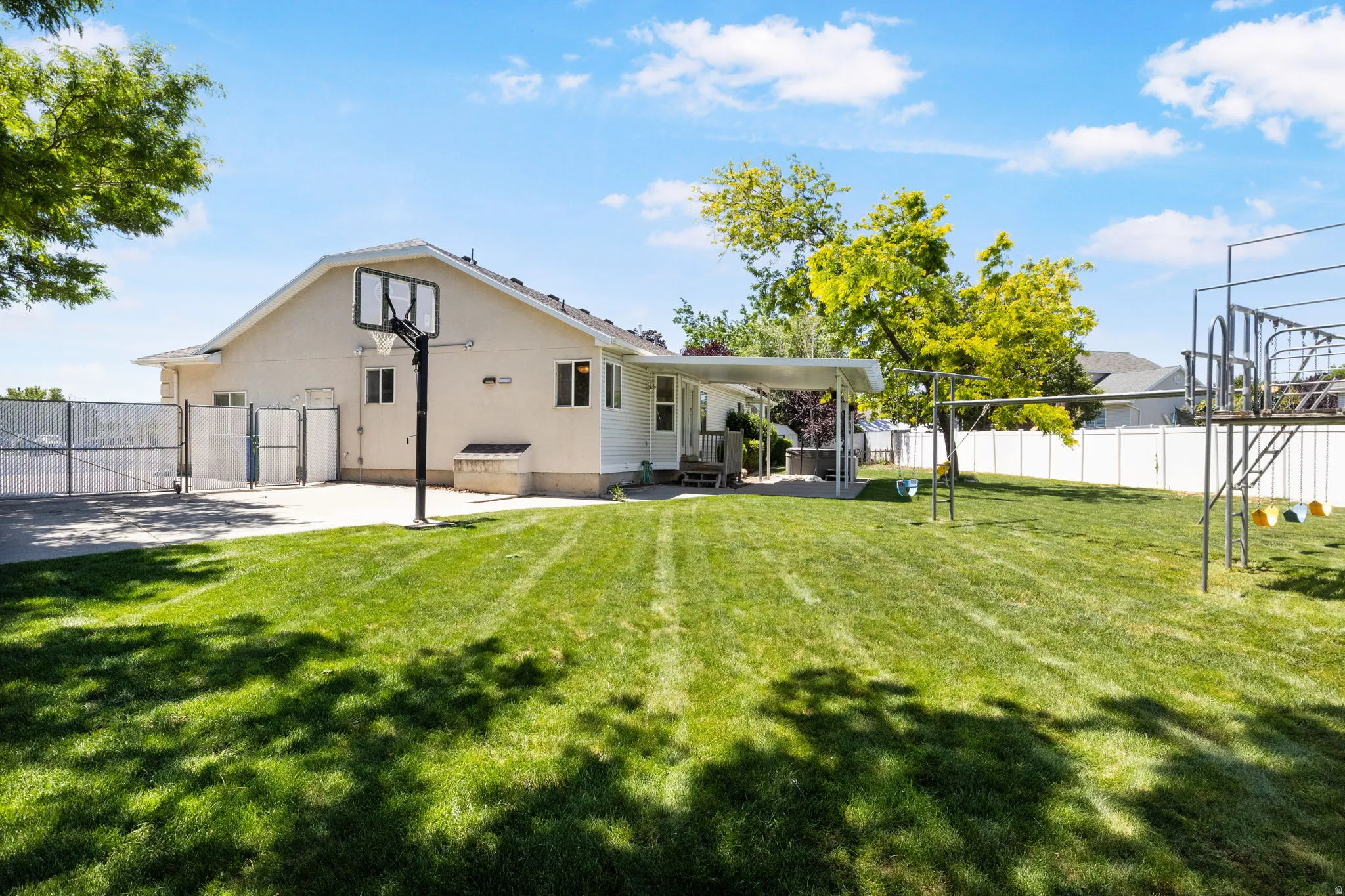 Expansive backyard featuring a large lawn area, full basketball hoop, white vinyl fencing, and a covered patio area