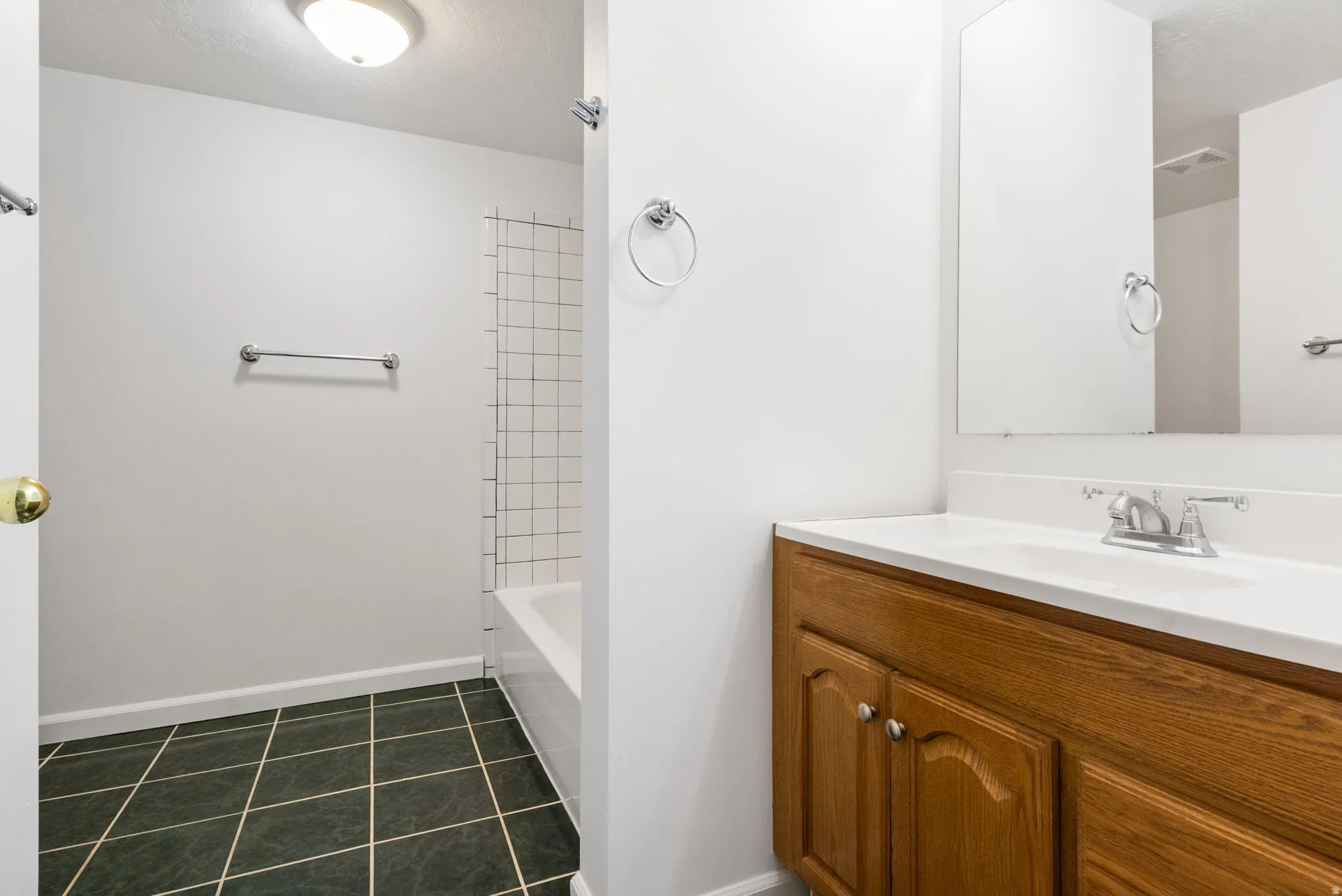 Bathroom featuring a white bathtub with tile surround, dark square tile flooring, and a wood-finish vanity with a white countertop and integrated sink