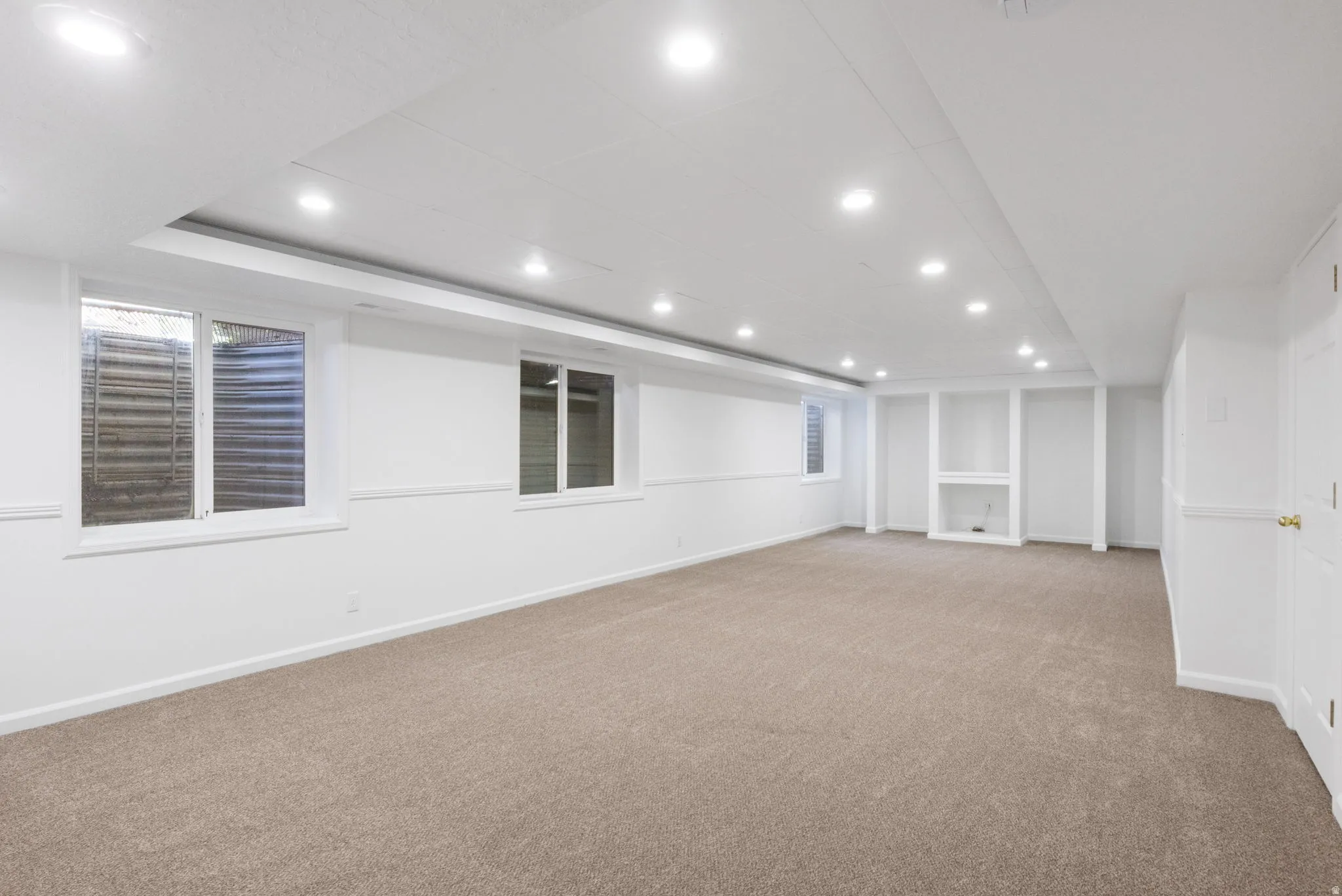 Spacious room featuring neutral carpeting, white walls with wainscoting, multiple recessed lights, and several windows for natural illumination
