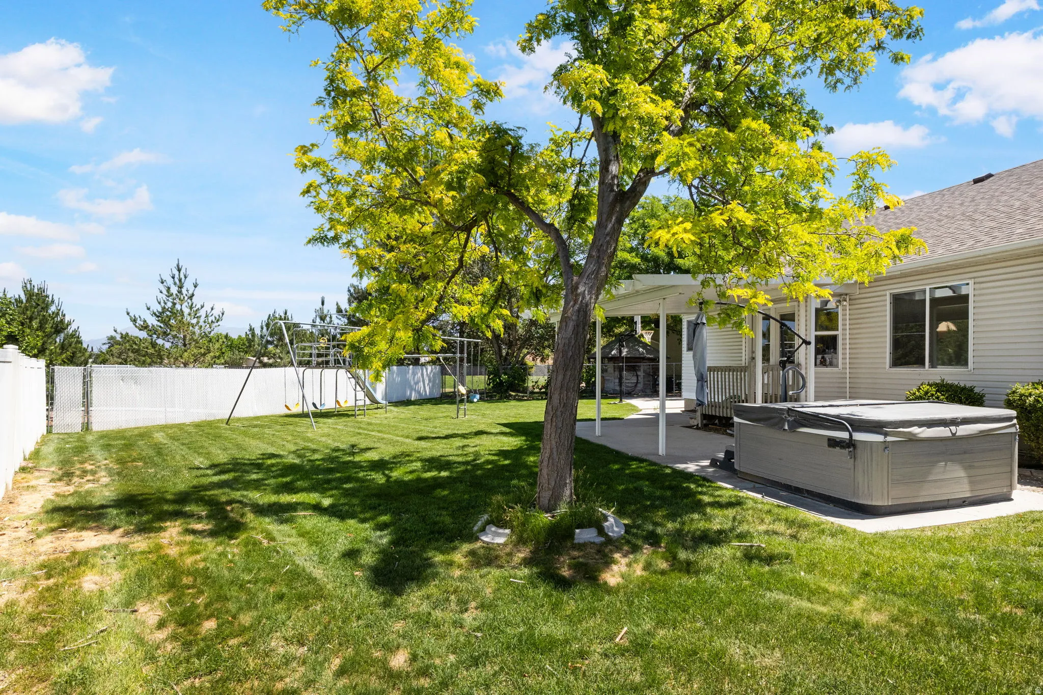 Expansive green lawn featuring a mature shade tree, a white vinyl privacy fence, a concrete patio, and a built-in hot tub