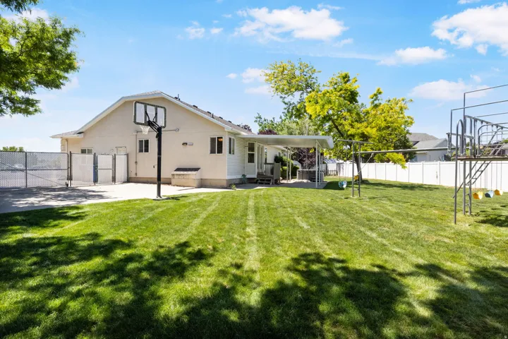 Expansive backyard featuring a large lawn area, full basketball hoop, white vinyl fencing, and a covered patio area