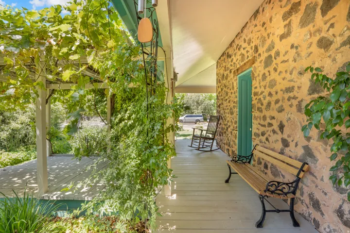 Inviting covered porch featuring a vine-covered pergola, plank flooring, a natural stone wall, and a vibrant teal entry door