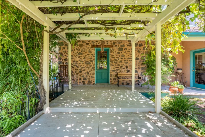Front porch with a white pergola featuring established vines, stone wall facade, teal entry door, and wood-finish decking