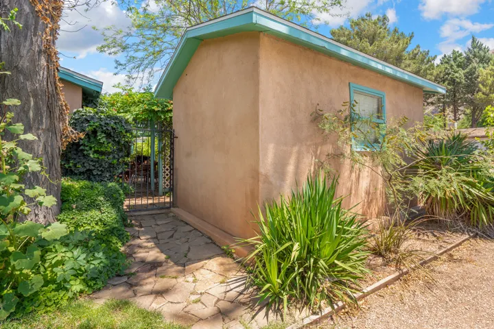 Stucco exterior with a teal trim window, featuring a stone path leading to a decorative iron gate, surrounded by mature trees and varied plantings