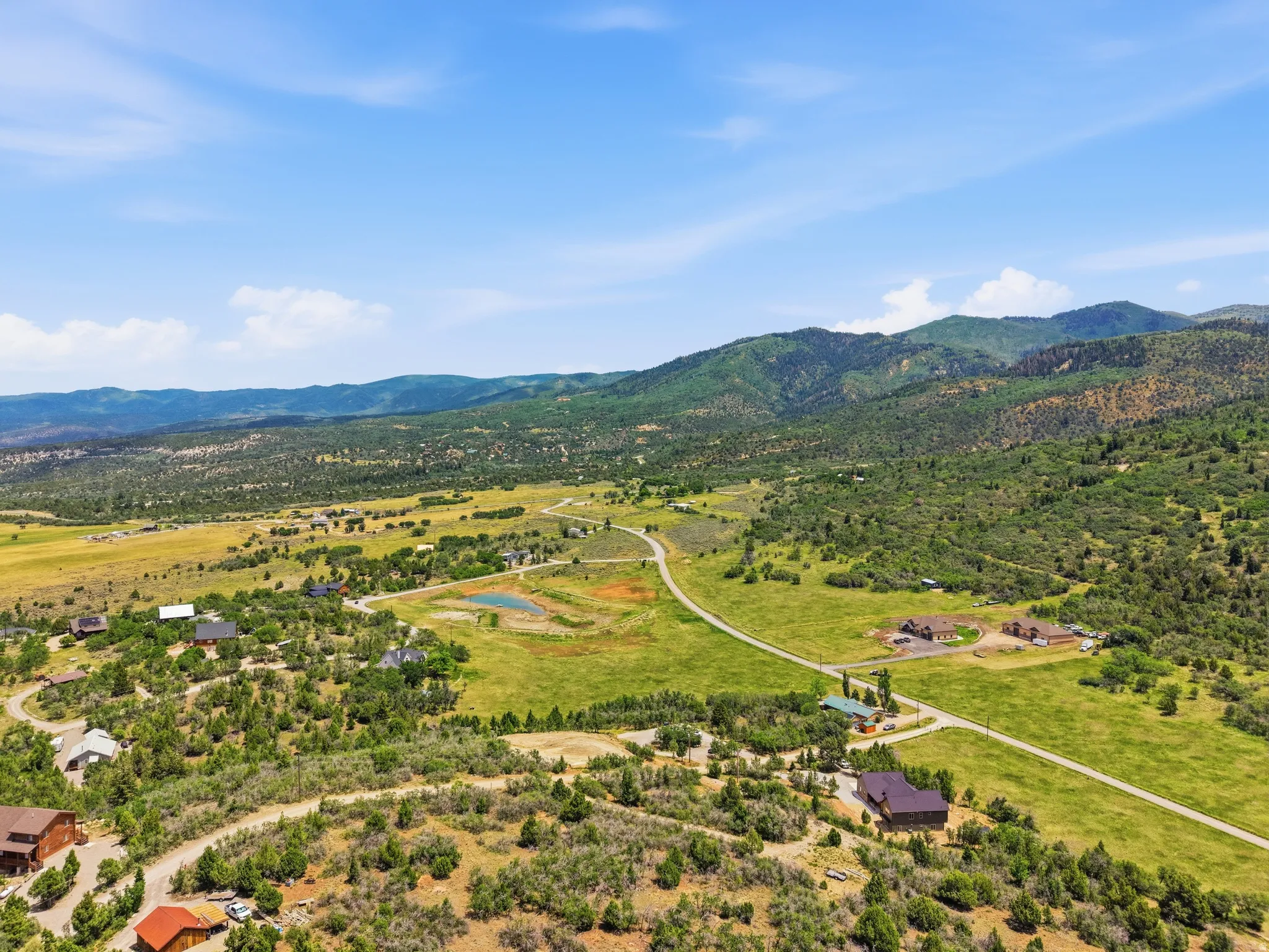 Expansive mountain landscape featuring rolling green hills, scattered residential structures, and a winding paved road