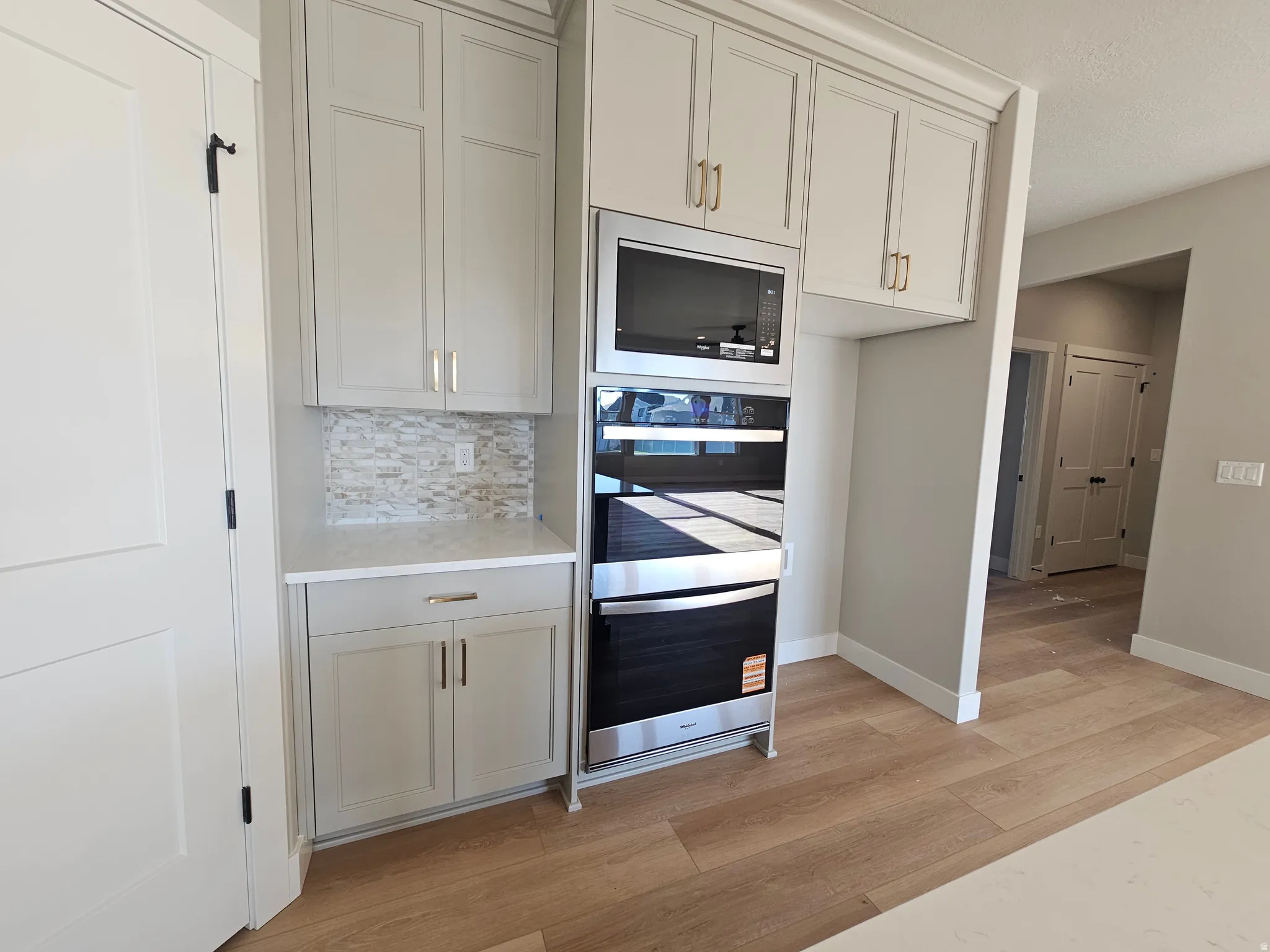 Built-in stainless steel microwave and oven, light grey cabinetry with gold-tone hardware, white countertop, stone mosaic tile backsplash, and light wood-finish flooring