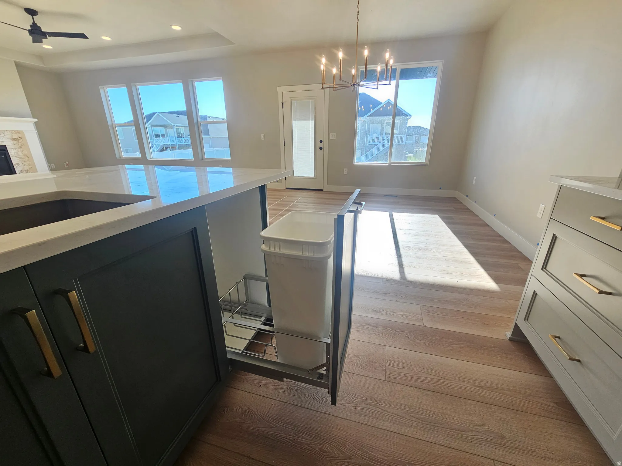 Open-concept interior featuring light wood-finish flooring, a contemporary chandelier, and a kitchen island with dark cabinetry, integrated sink, and pull-out waste bin