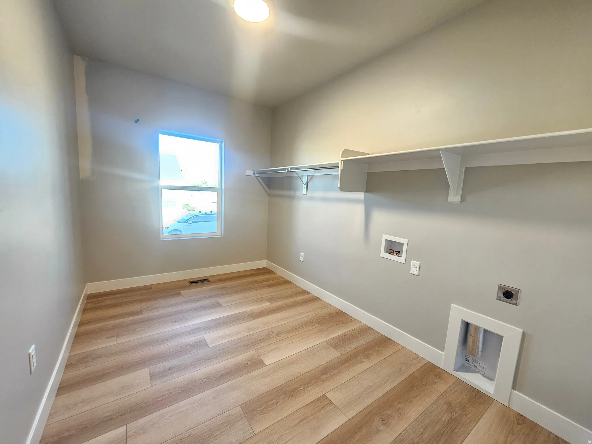 Laundry room featuring wood-finish flooring, a single window, overhead built-in shelving, and white baseboards