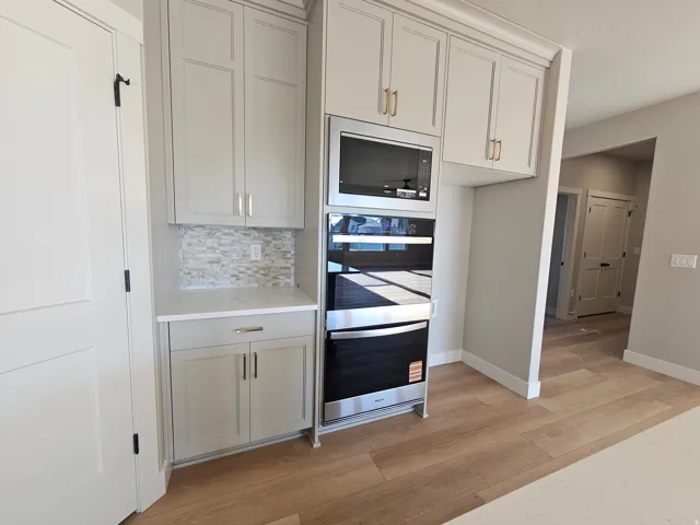 Built-in stainless steel microwave and oven, light grey cabinetry with gold-tone hardware, white countertop, stone mosaic tile backsplash, and light wood-finish flooring