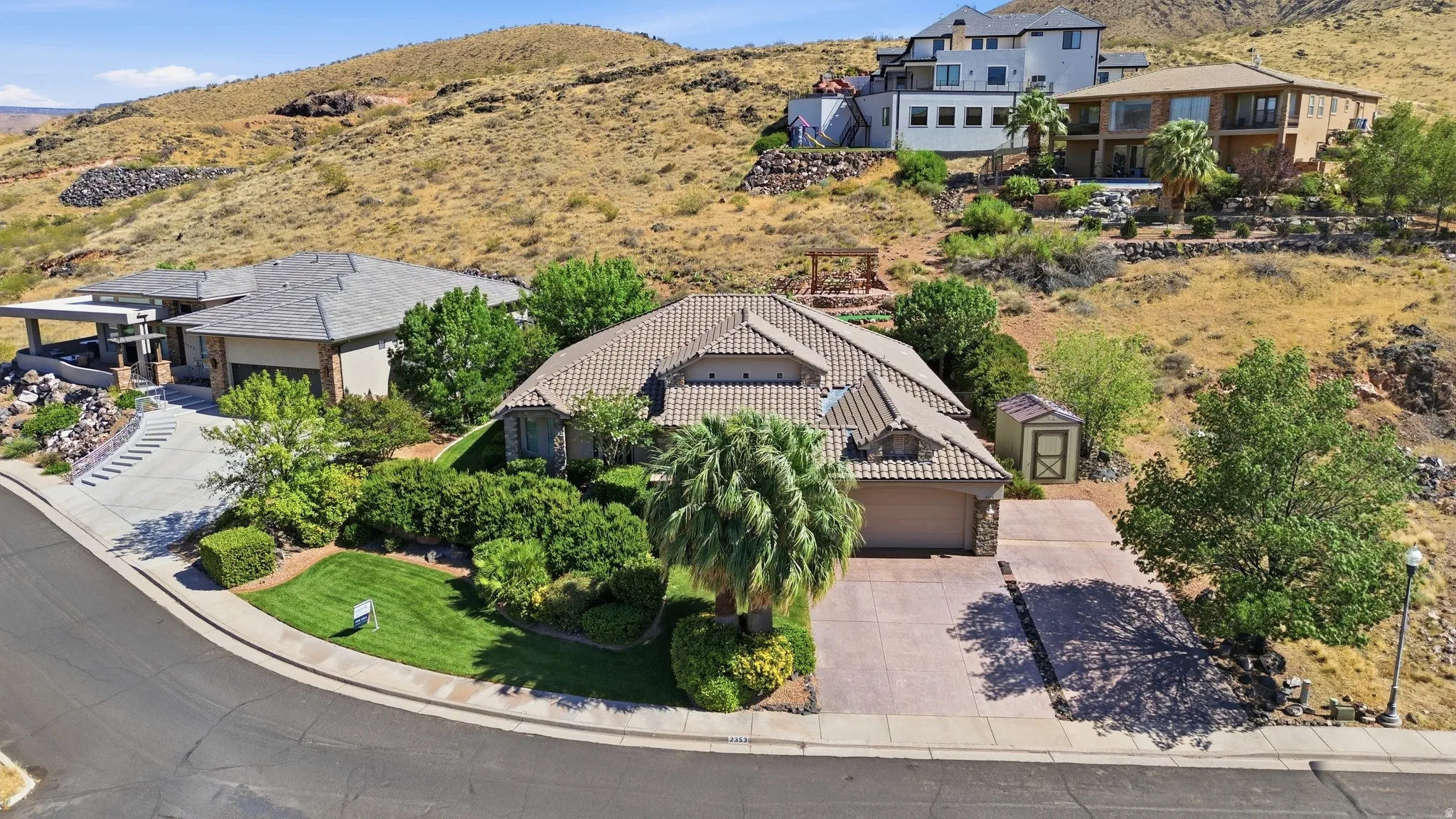 Terracotta tile roof home featuring a two-car garage, mature landscaping, paved driveway, and a storage shed