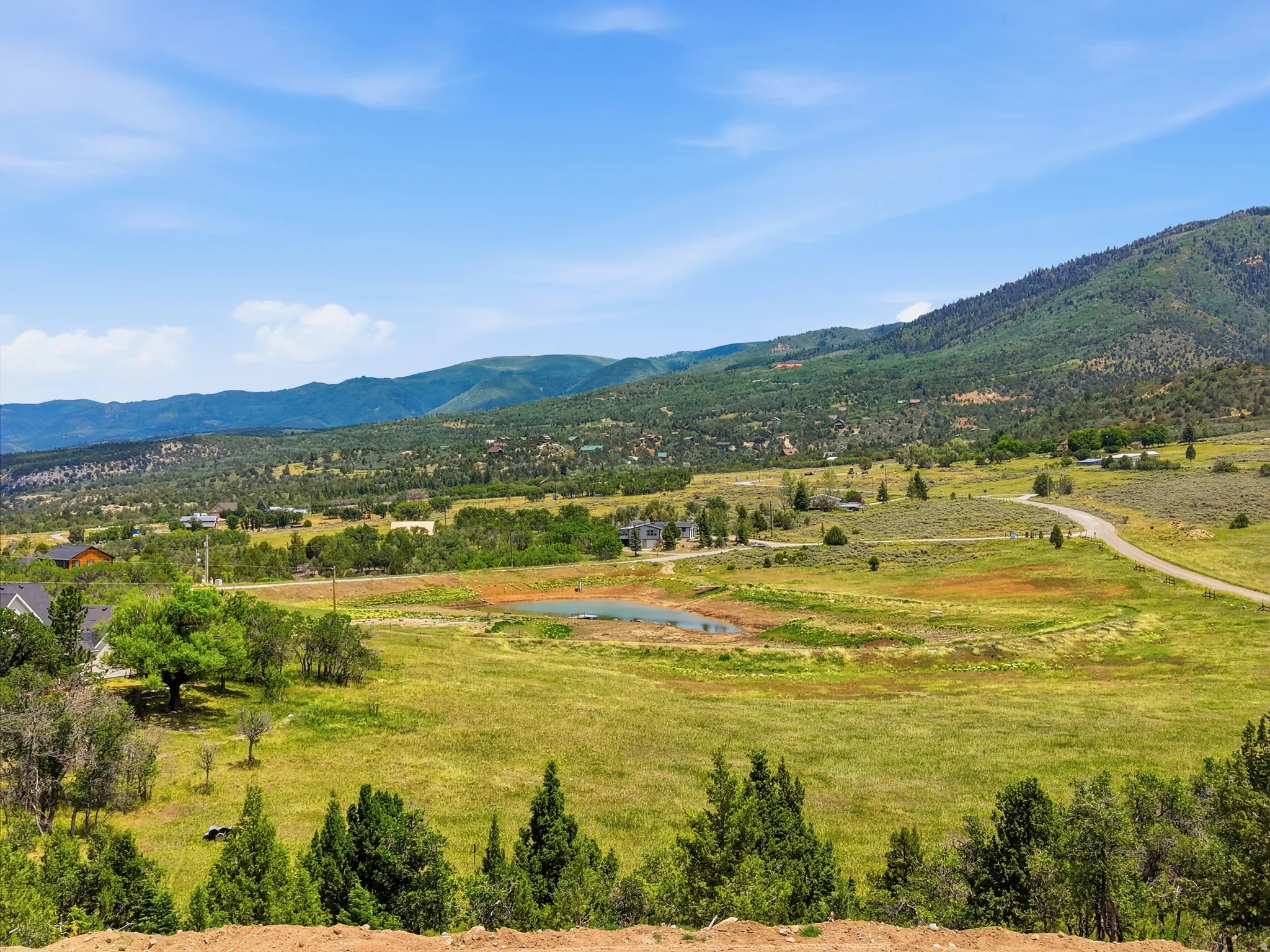 Expansive land parcel featuring a natural pond, a paved road, scattered mature trees, and mountainous terrain in the background