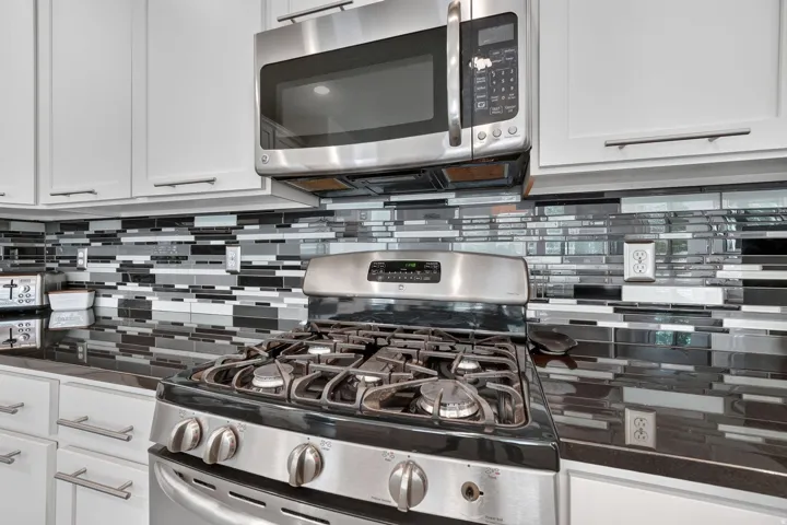 Stainless steel gas range and microwave oven paired with white cabinetry, dark countertops, and a mosaic tile backsplash