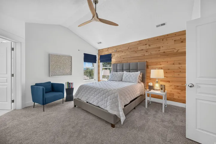 Vaulted ceiling bedroom featuring a natural wood accent wall, two windows with navy Roman blinds, and light grey carpet flooring