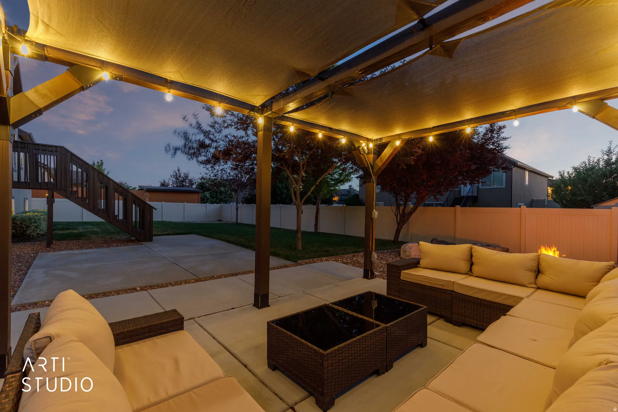 Covered patio featuring string lighting, concrete pavers, a privacy fence, and a staircase with wood railings