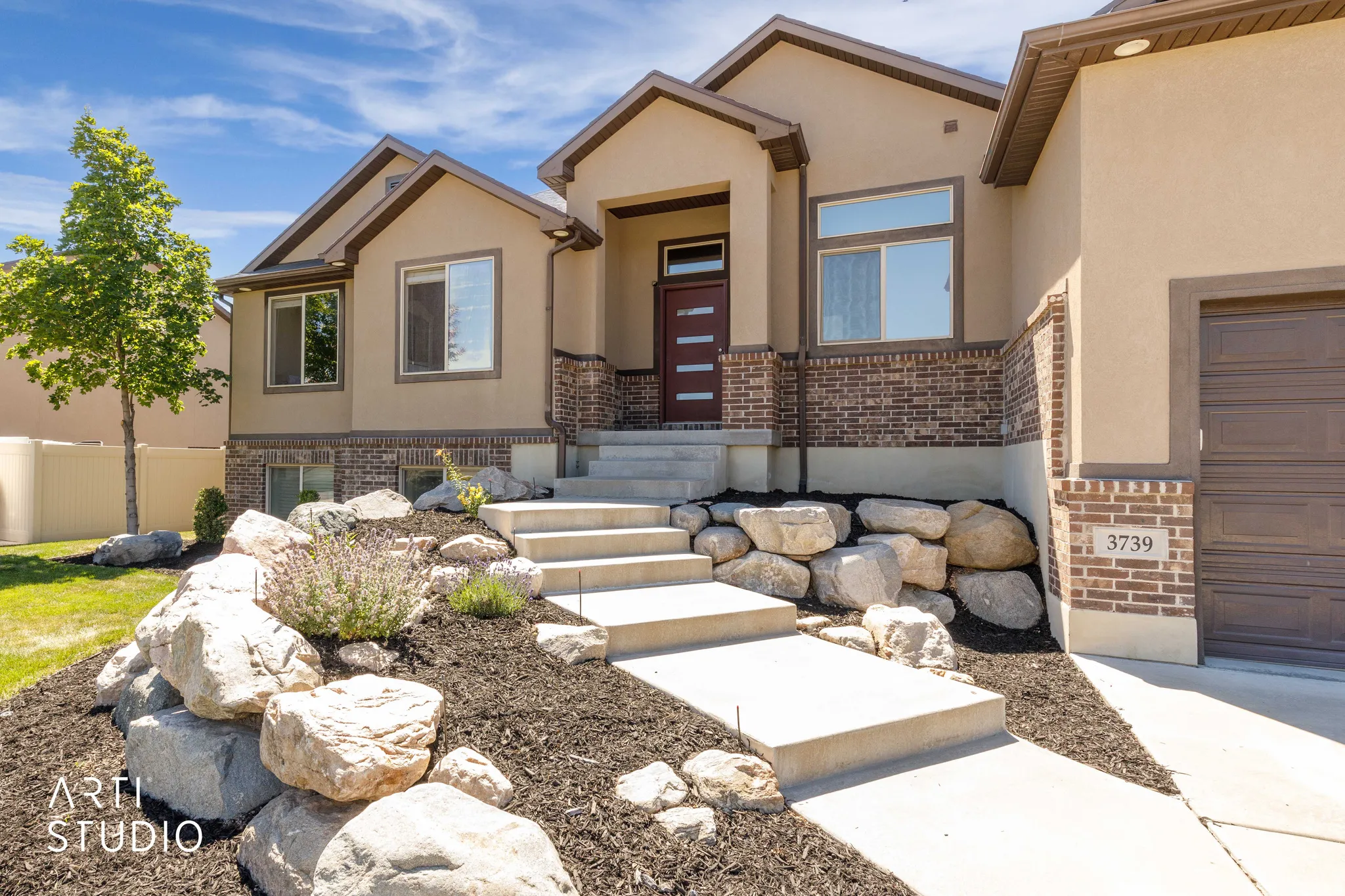 Contemporary stucco and brick exterior featuring a multi-paneled entry door, concrete walkway, tiered landscaping with natural boulders, and an integrated garage